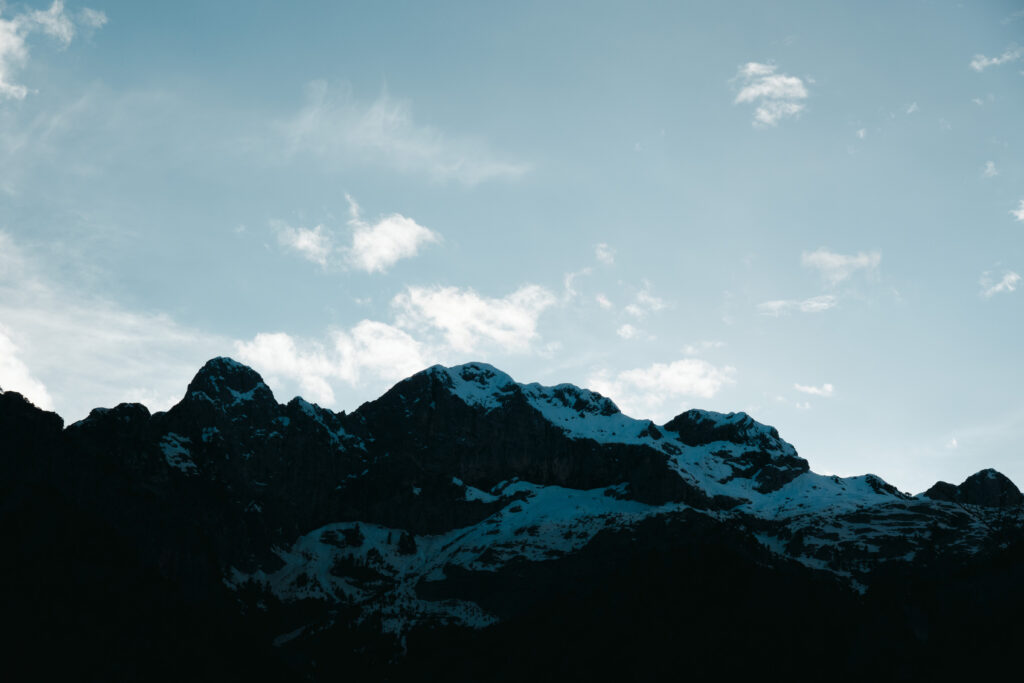 A wide landscape view of a towering mountain range, predominantly silhouetted in dark, deep blues and blacks, with several prominent peaks partially covered in white and light blue snow. The upper half of the image features a clear, pale blue sky with scattered, wispy white clouds. The dramatic contrast highlights the rugged terrain and the vastness of the natural environment.