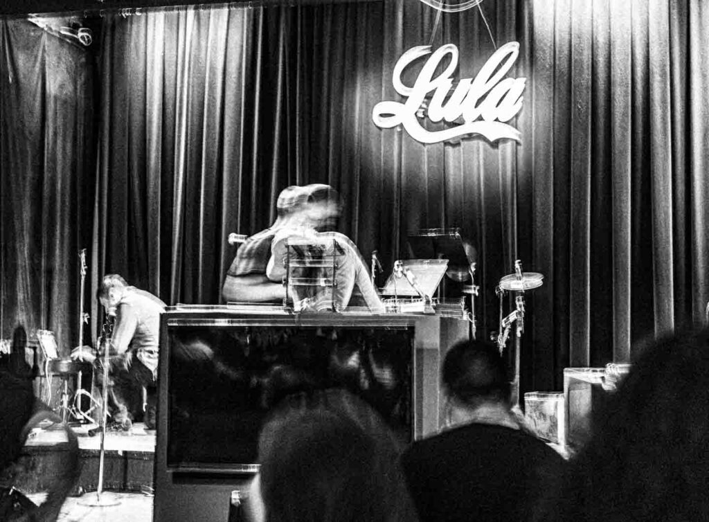 A stark black and white, grainy photograph captures a scene from a live music event, characterized by significant motion blur. Dominating the upper center is a bright, cursive 'Lula' sign against dark, pleated stage curtains. On the stage floor, a musician on the left bends over equipment next to a microphone stand. In the center-right, two highly blurred figures, likely musicians, are positioned behind a reflective piece of equipment, possibly a large console or piano, with elements of a drum kit visible further right. The foreground shows the indistinct, dark forms of audience members from behind, watching the blurred activity on stage, conveying a sense of movement and immersion in the performance.