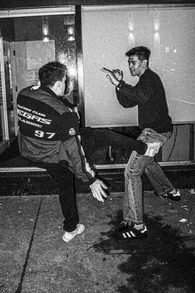 A grainy black and white photograph captures a candid street scene at night featuring two men. On the left, a man with dark curly hair, seen from the side, wears a black and red 'RACING CLUB VEGAS CLASSIC 97' jacket. He is sitting on a dark ledge, his right arm extended with an open hand towards the ground. On the right, a man with short dark hair and a faint smile is caught mid-action, appearing to jump or dance. He wears a dark long-sleeved shirt, distressed jeans, and dark sneakers with white stripes. He holds a cigarette between the fingers of his uplifted left hand, while his right hand is also open and raised. The legs of a third person are faintly visible between them. The background includes a reflective storefront window and a light-colored wall. The concrete sidewalk is speckled with debris and reflects light in wet patches.