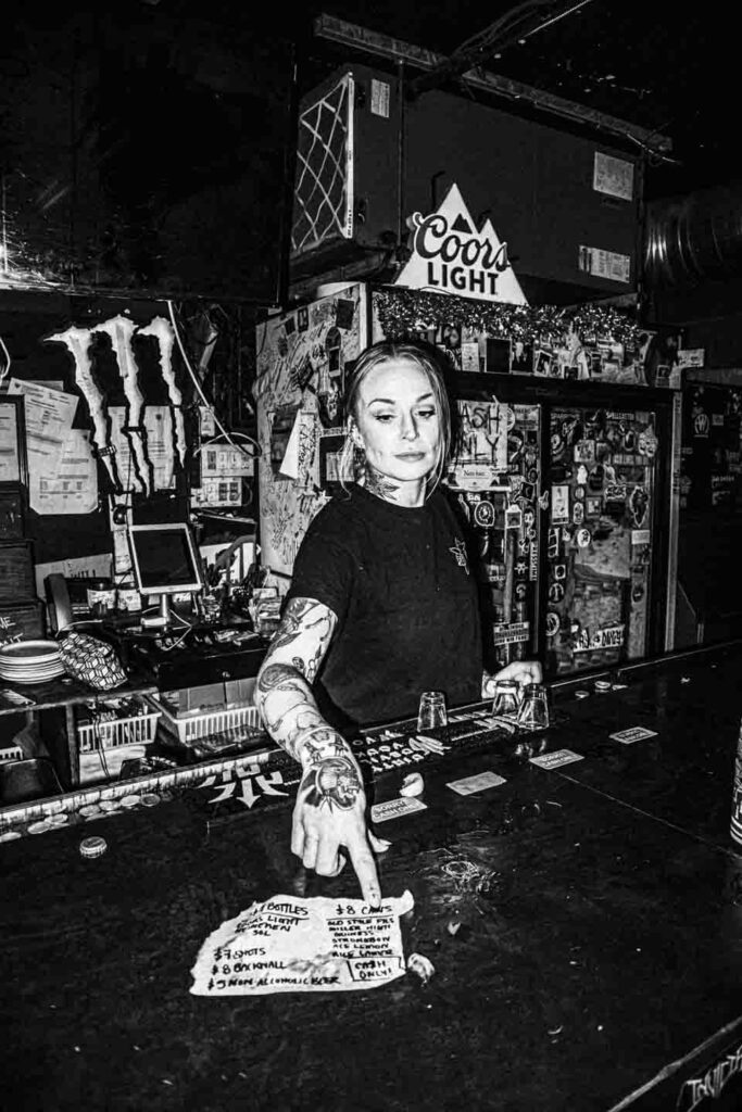 A black and white image of a young female bartender with extensive arm and neck tattoos, wearing a dark t-shirt, standing behind a dimly lit bar. She is looking down and pointing her right finger at a crumpled, handwritten paper menu on the sticky bar surface that includes 'CASH ONLY!'. The background features a cluttered wall covered in stickers and posters, a large Monster Energy logo on the left, and a prominent Coors Light sign hanging above.