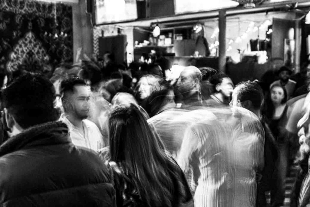 A black and white photo captures a dense crowd of people in a bustling indoor space, likely a bar or club. Many figures are rendered with significant motion blur, especially in the mid-ground and background, creating a sense of dynamic activity. In the foreground, the backs of people's heads and shoulders are visible, with one man's bearded profile slightly clearer amidst the movement. Overhead, indistinct architectural details and lighting fixtures suggest a vibrant social environment.