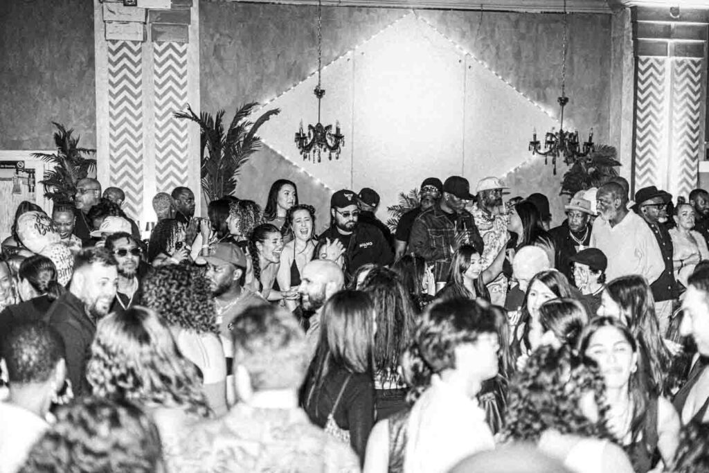 A black and white photograph depicting a large crowd of people packed into a dimly lit indoor venue. Many attendees are smiling and interacting, with a 'Happy Birth' balloon visible on the left. The background features ornate walls with chevron-patterned pillars, a large diamond-shaped light display with string lights, two chandeliers, and potted palm plants, creating an energetic party atmosphere.