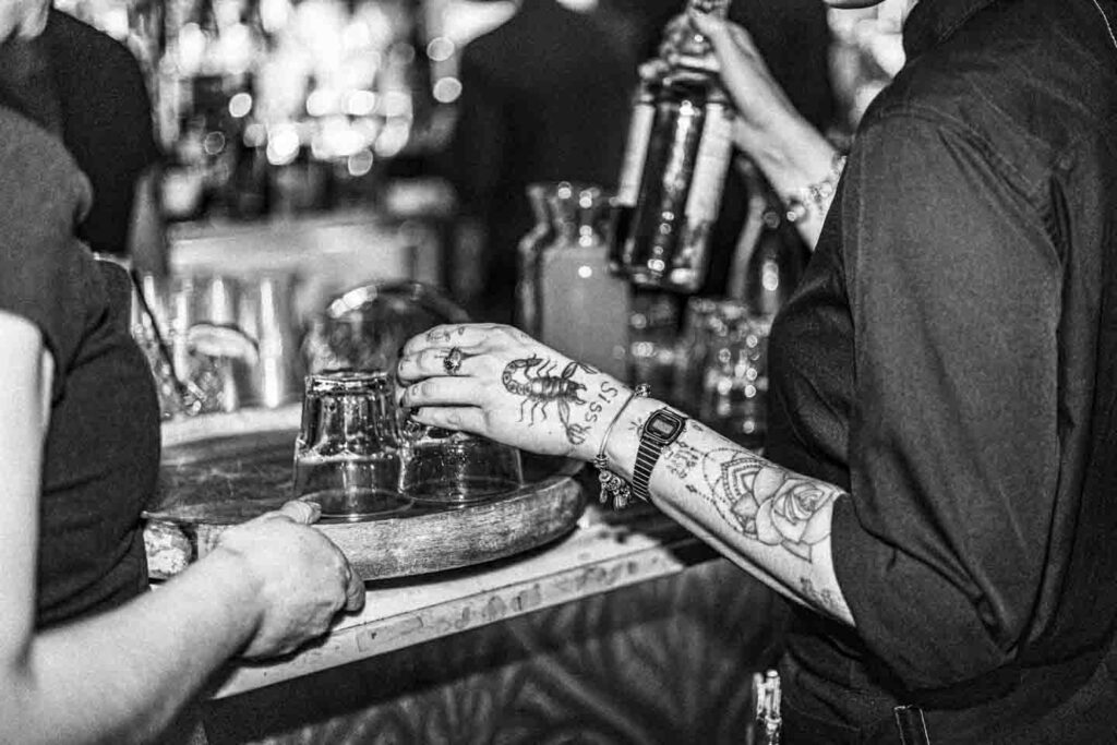 A black and white, close-up shot of a bustling bar. On the right, a person's tattooed arm, wearing a Casio digital watch and rings, holds a liquor bottle. Tattoos include a scorpion, the word 'SISSY,' and various ornamental patterns. On the left, another person's hands rest on a round wooden tray containing two empty drinking glasses. The background is blurred with bar lights, bottles, and other glassware.