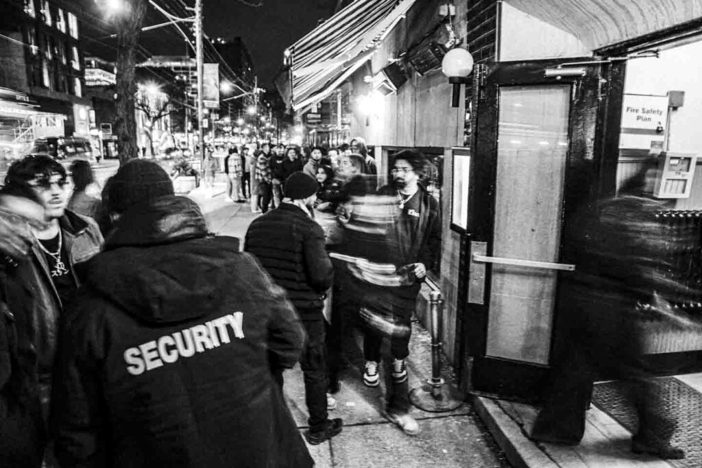 A high-contrast black and white photograph captures a busy nighttime street scene. In the foreground, a person wearing a jacket with 'SECURITY' on the back stands with their back to the viewer. To their right, an open doorway leads into a building, with a 'Fire Safety Plan' sign visible. Several people are gathered near the entrance, some with motion blur indicating movement, while a line of people extends down the sidewalk under the illumination of distant streetlights.