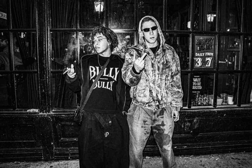 A black and white street photograph depicts two young men standing in front of a dark, windowed building. The man on the left has curly hair and glasses, wearing a 'BULLY' shirt and black baggy pants, holding a lit cigarette and exhaling smoke. The man on the right wears a camouflage hooded jacket and sunglasses, with a cigarette in his mouth, raising his right hand to show the middle finger. Behind them, 'HAPPY HOUR' and 'PLEASE WAIT TO BE SEATED' signs are visible in the windows.