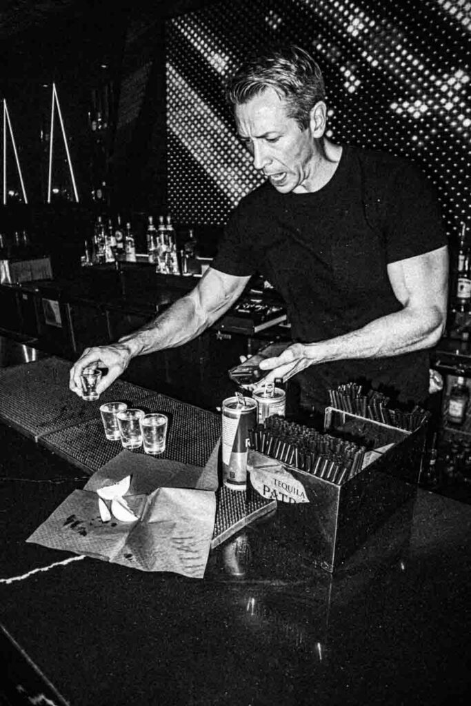 A high-contrast black and white shot of a male bartender with short, light hair and a black t-shirt, actively preparing drinks at a bar. He holds one shot glass in his left hand over three others on a rubber mat. On the counter are lime wedges on a crumpled napkin, two beverage cans, and a Tequila Patrón branded dispenser full of stirrers. Bottles line the shelves behind him, and a textured, possibly illuminated, wall fills the upper background.
