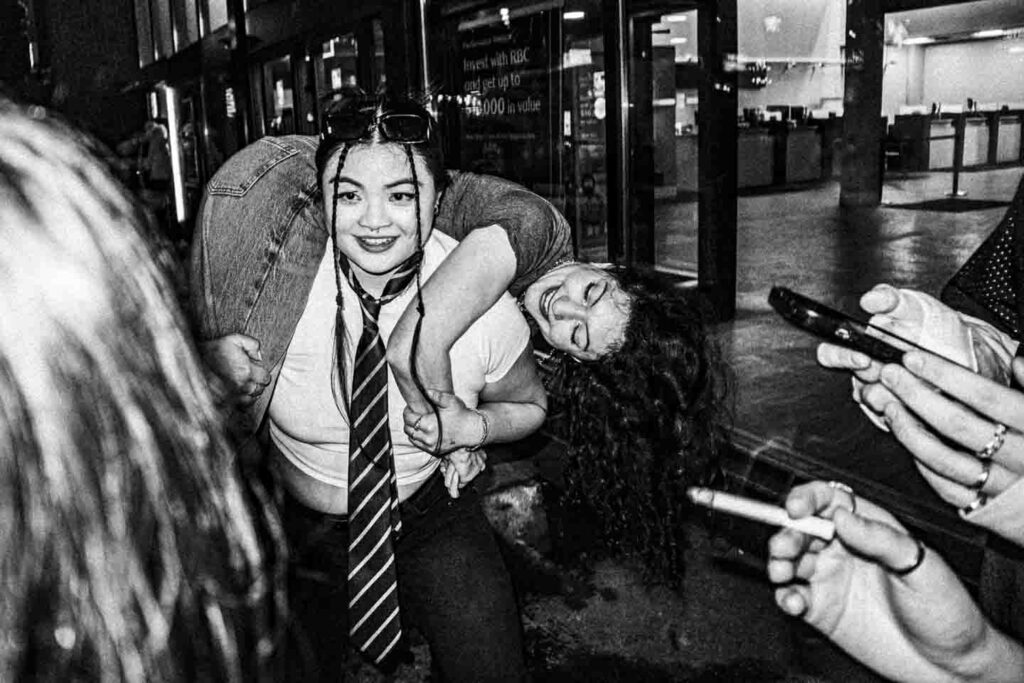 A grainy, black and white photograph features two young women: one with braided hair and a striped tie smiles at the camera while giving a piggyback ride to another woman with long, dark curly hair, who is laughing with her eyes closed. They are indoors, with a sign in the background reading 'Invest with RBC.' In the lower right foreground, hands are visible, one holding a smartphone and another a cigarette.