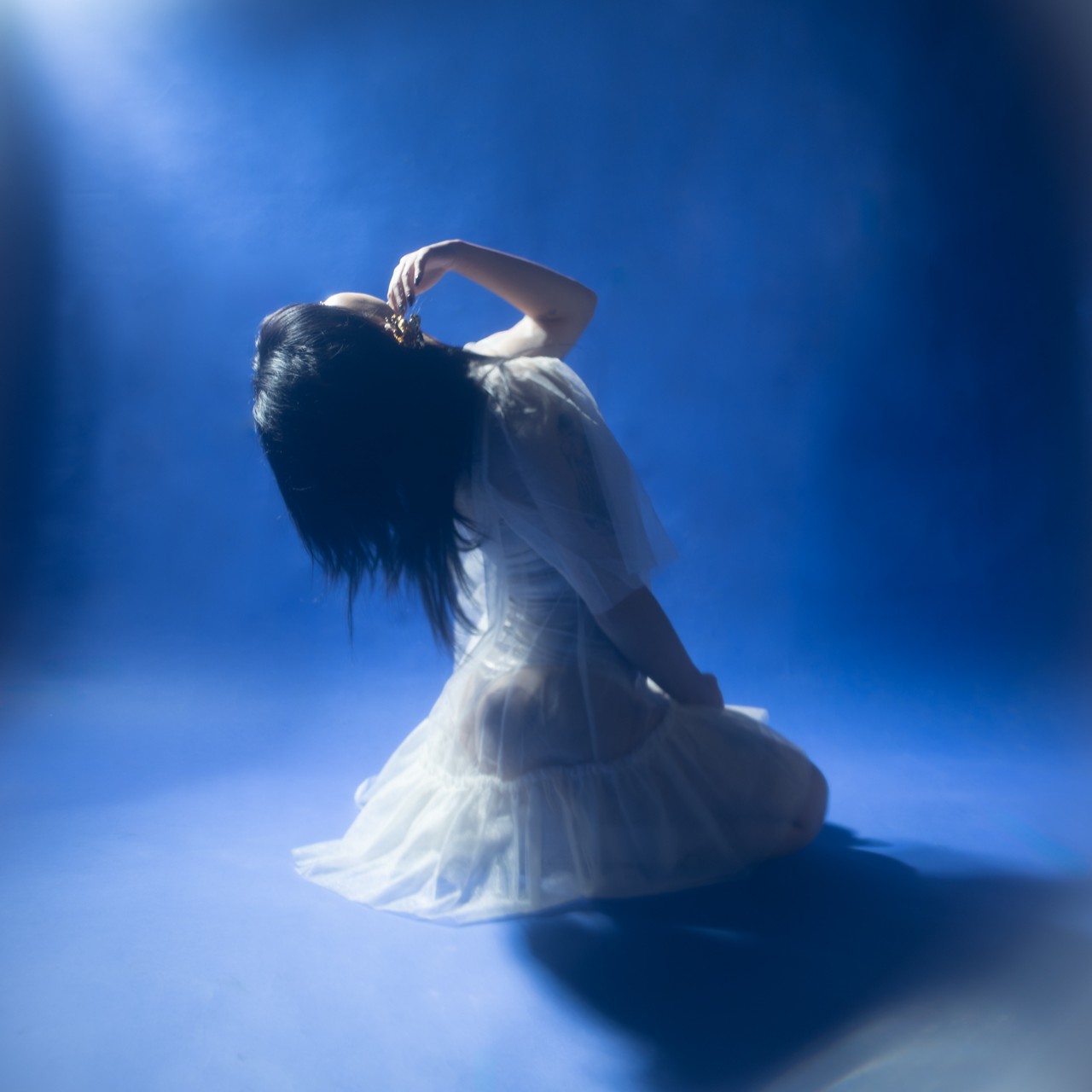 A full-body shot of a person with long dark flowing hair wearing a translucent white dress kneeling on a vibrant blue surface Their head is sharply tilted backward looking upwards as one hand delicately touches a golden intricate ornament near their temple A strong ethereal light source from the upper left illuminates the scene creating a pronounced dark shadow beneath the figure and a reflective glow on the blue ground lending an otherworldly and contemplative atmosphere