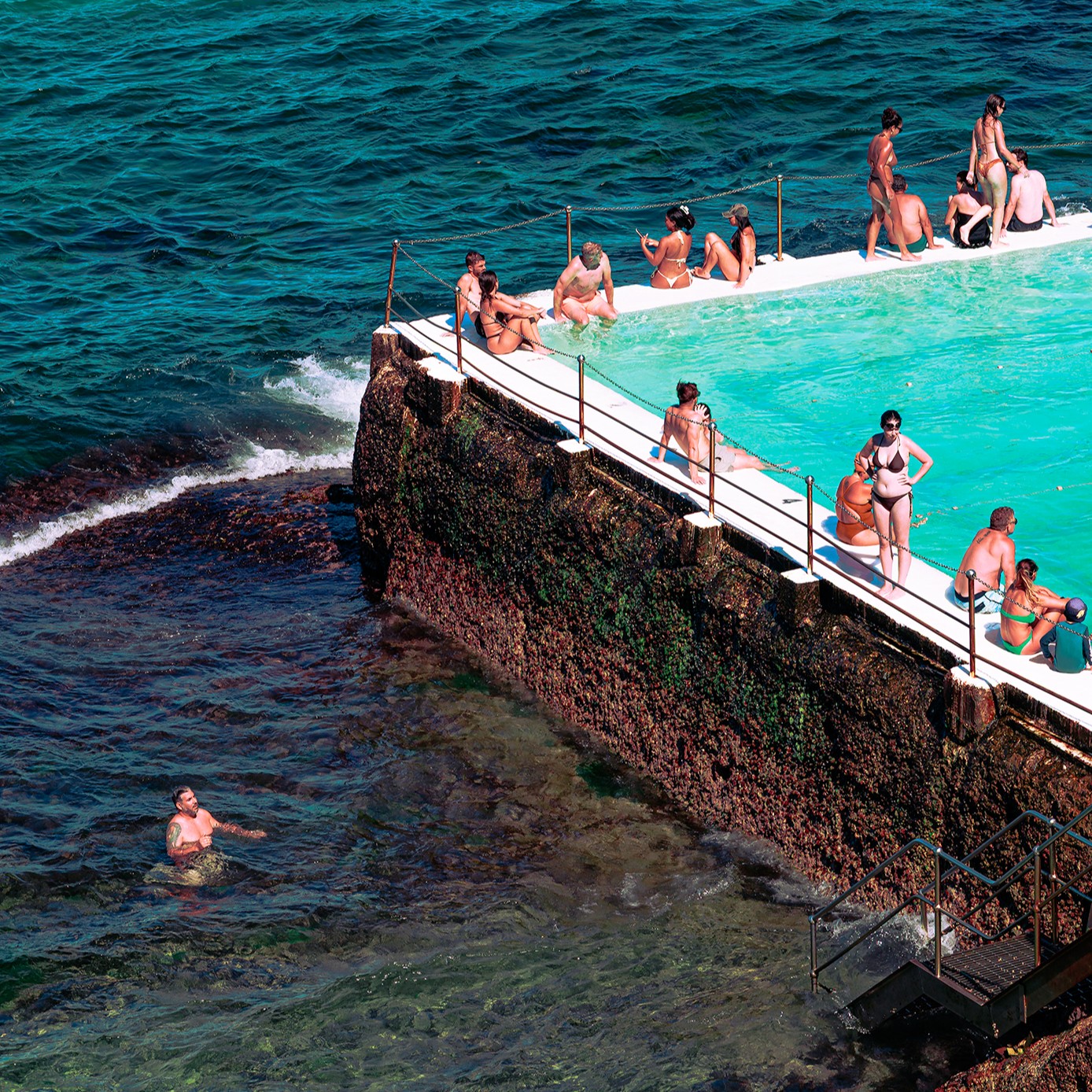 A high-angle view of a sunny coastal scene shows an ocean rock pool with vibrant turquoise water bordered by a white edge and chain railing Many people in swimwear are sitting on the edge or in the pool A rugged dark brown wall covered in texture separates the calm pool from the deep blue-green ocean with gentle waves A man with visible tattoos is wading in the ocean water to the left of the pool and metal steps lead into the sea on the right